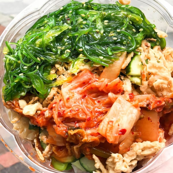 A vibrant Hawaiian poke bowl with green seaweed salad, red kimchi, and crispy toppings.